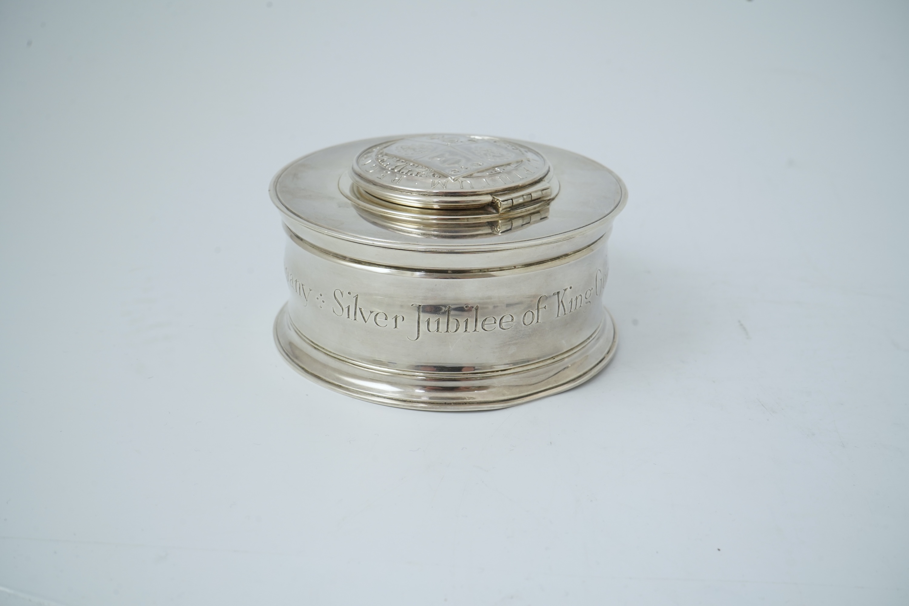 A George V silver circular inkwell, by Sebastian Garrard, commemorating the Silver Jubilee of King George V, the die stamped cover designed by George Kruger Gray featuring the Goldsmiths Company Coat of Arms